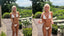 Woman with blonde braids wearing a white ruffled thong bikini, standing outdoors in a garden setting with greenery and flowers in the background.